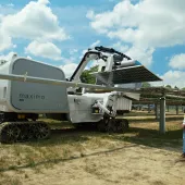 A robotic machine labeled 'maximo aes' is operating in a solar farm, adjusting a solar panel. The machine is on caterpillar tracks, and a person in a safety vest and helmet observes nearby. The background shows rows of solar panels under a partly cloudy sky.