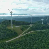 Aerial view of a lush green landscape with several wind turbines on hilltops. A winding road passes through the hills, and clouds hover over distant mountains under a partly cloudy sky.