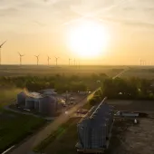 Aerial view of a rural landscape at sunset with wind turbines in the distance. The foreground features several large silos and farm buildings, with trees and open fields surrounding them. The sky is filled with a warm golden hue from the setting sun.