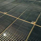 Aerial view of a large solar farm with multiple rows of solar panels set in a grassy field. The panels are aligned in parallel rows, surrounded by green trees and open fields in the background.