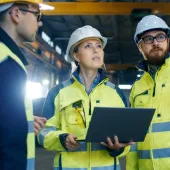 Three construction workers wearing safety helmets and high-visibility jackets stand inside a large industrial facility. One holds a laptop, and they appear to be discussing or inspecting something above.