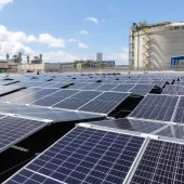 A large field of solar panels under a blue sky, with an industrial facility and a large cylindrical structure in the background.