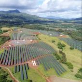 Aerial view of a large solar farm with rows of solar panels spread across a green landscape. Mountains are visible in the background under a partly cloudy sky.