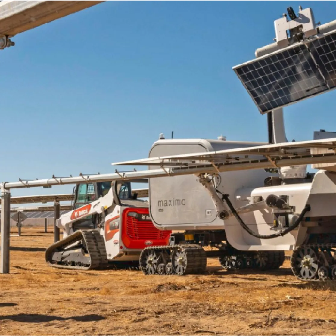 Two workers in safety vests and helmets observe a robotic machine cleaning solar panels in a solar farm under a clear blue sky.