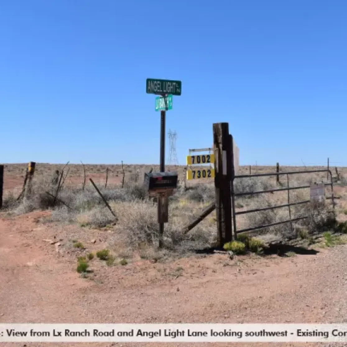 Intersection of Lx Ranch Road and Angel Light Lane in a rural area, looking southwest. Dirt roads intersect with a metal gate on the left and a fence on the right. Road signs display 'Angel Light Ln' and 'Lx Ranch Rd'. Sparse vegetation and clear blue sky are visible.