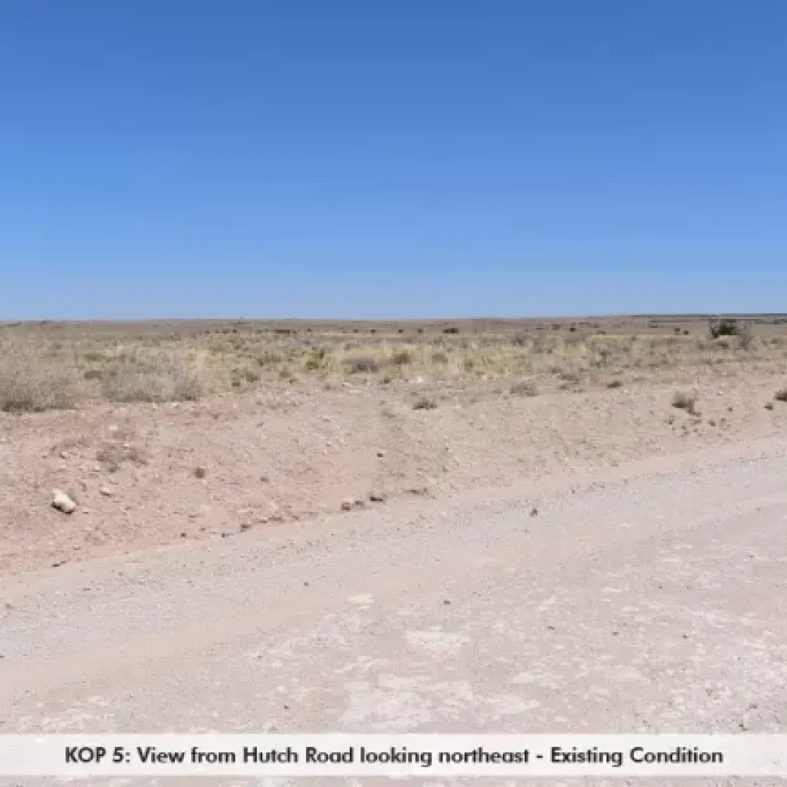 A dry, barren landscape viewed from Hutch Road looking northeast. The foreground shows a dirt road with sparse vegetation on either side. The background features a flat, open expanse under a clear blue sky.