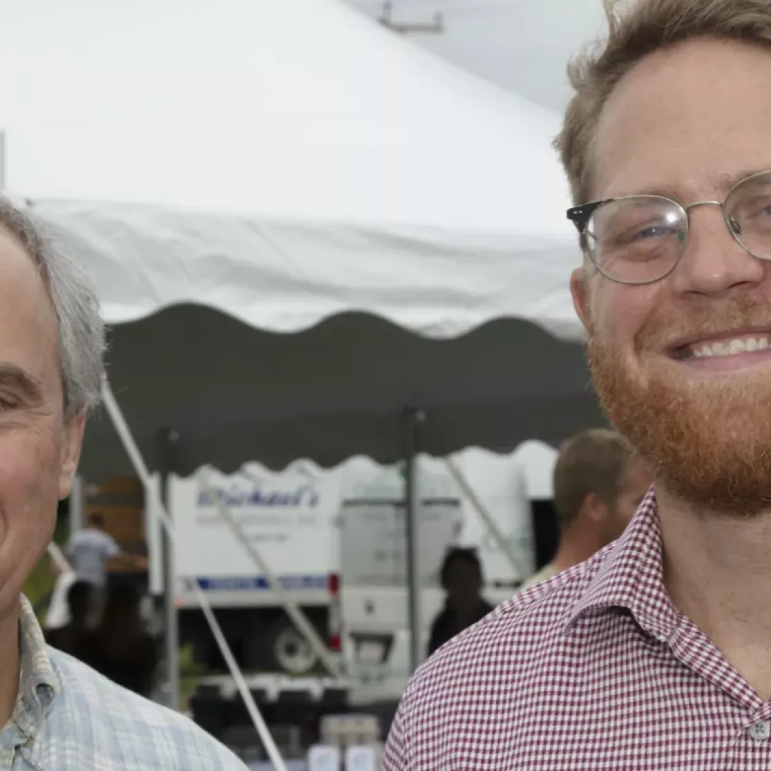 Two men smiling at an outdoor event with a white tent in the background. The man on the left has gray hair and is wearing a plaid shirt, while the man on the right has red hair, a beard, glasses, and is wearing a checkered shirt. Other people and trees are visible in the background.