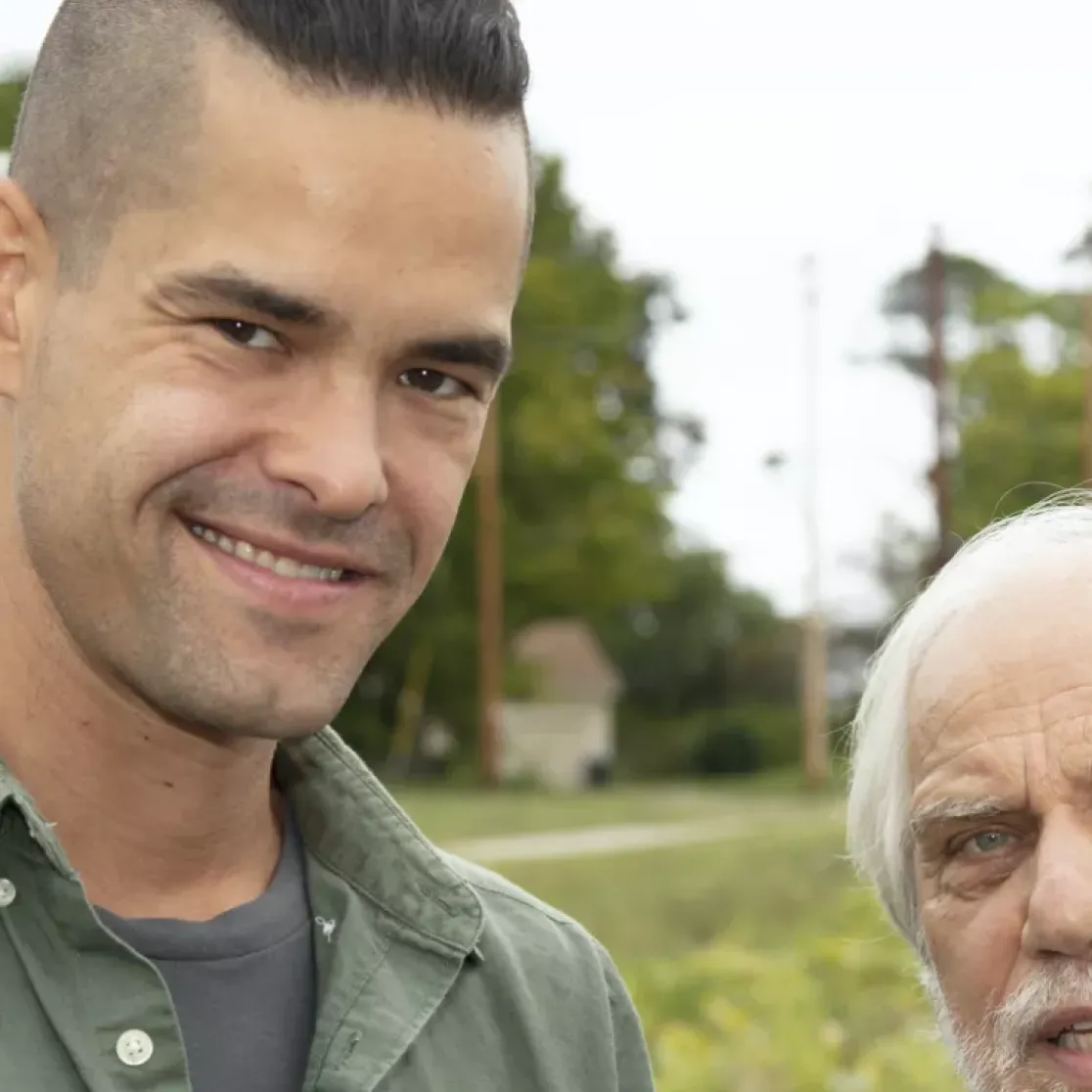Two men are standing outdoors in a field with trees in the background. The man on the left has short dark hair and is wearing a green shirt. The man on the right has white hair and a beard, wearing a light-colored shirt. Both are smiling softly at the camera.