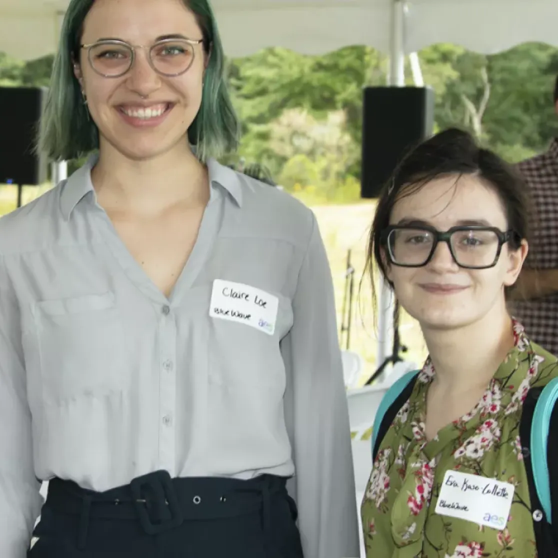 Three people smiling at the camera under a tent during an outdoor event. They are wearing name tags. In the background, a group of people are conversing and a projector screen is visible.