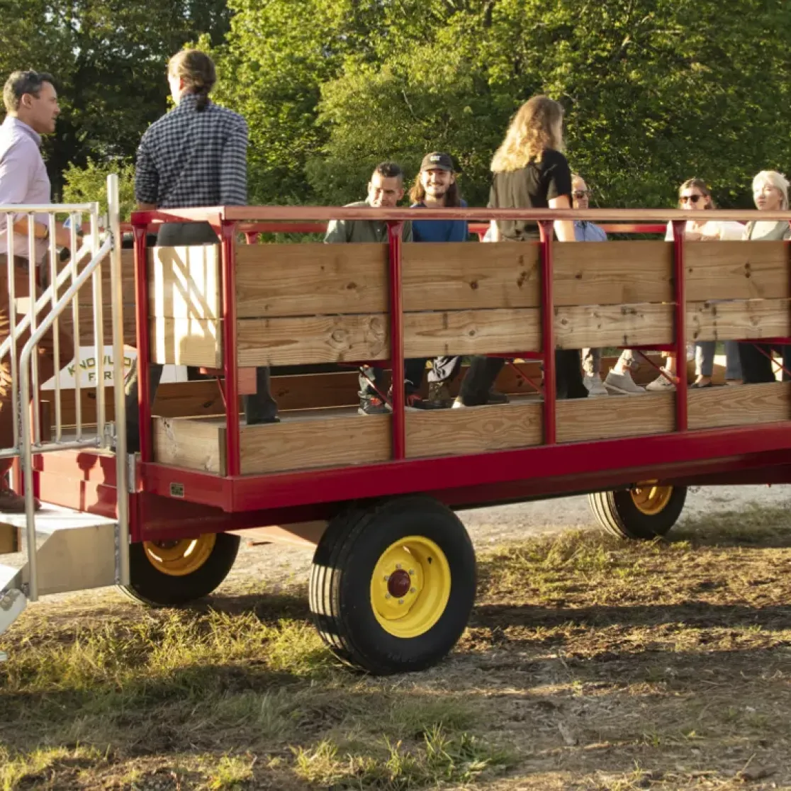 A group of people boarding a wooden wagon attached to a green tractor in a rural setting. Some are climbing stairs onto the wagon, while others are already seated. Trees and a dirt path are visible in the background.
