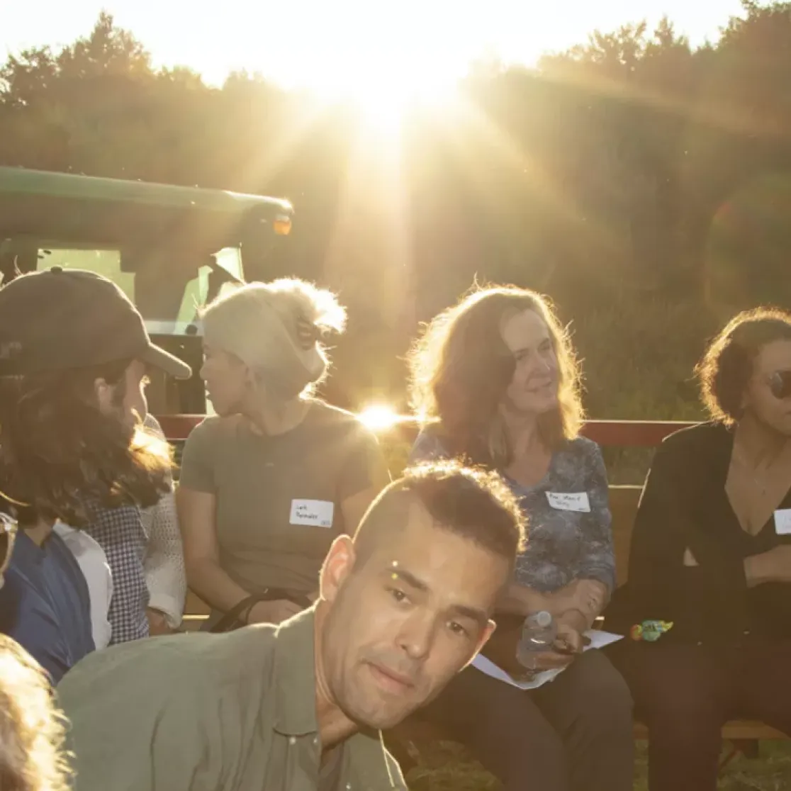 A group of people sitting outdoors in a circle, engaged in conversation. The sun is setting in the background, casting a warm glow. A green tractor is visible to the left. The group is diverse in age and gender, and some are wearing name tags.