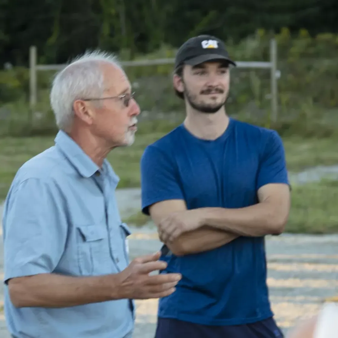 A group of people gathered outdoors in a conversation. An older man in a light blue shirt is speaking, while a younger man in a blue T-shirt with crossed arms listens. Others are partially visible, with trees and grass in the background.