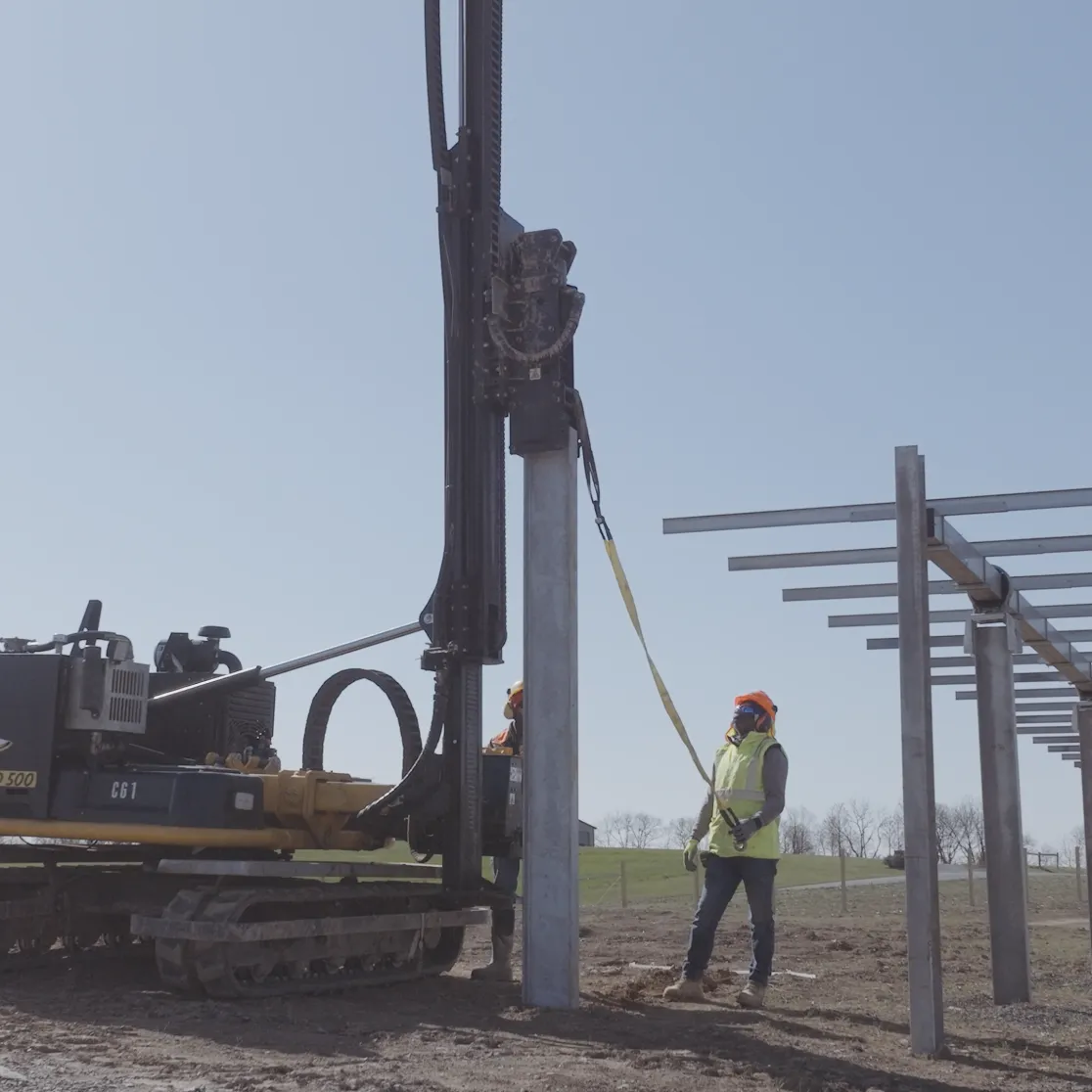 A construction worker in a safety vest and helmet stands next to a pile driver machine installing metal posts in a field. The posts form a long row extending into the distance. The sky is clear and blue.