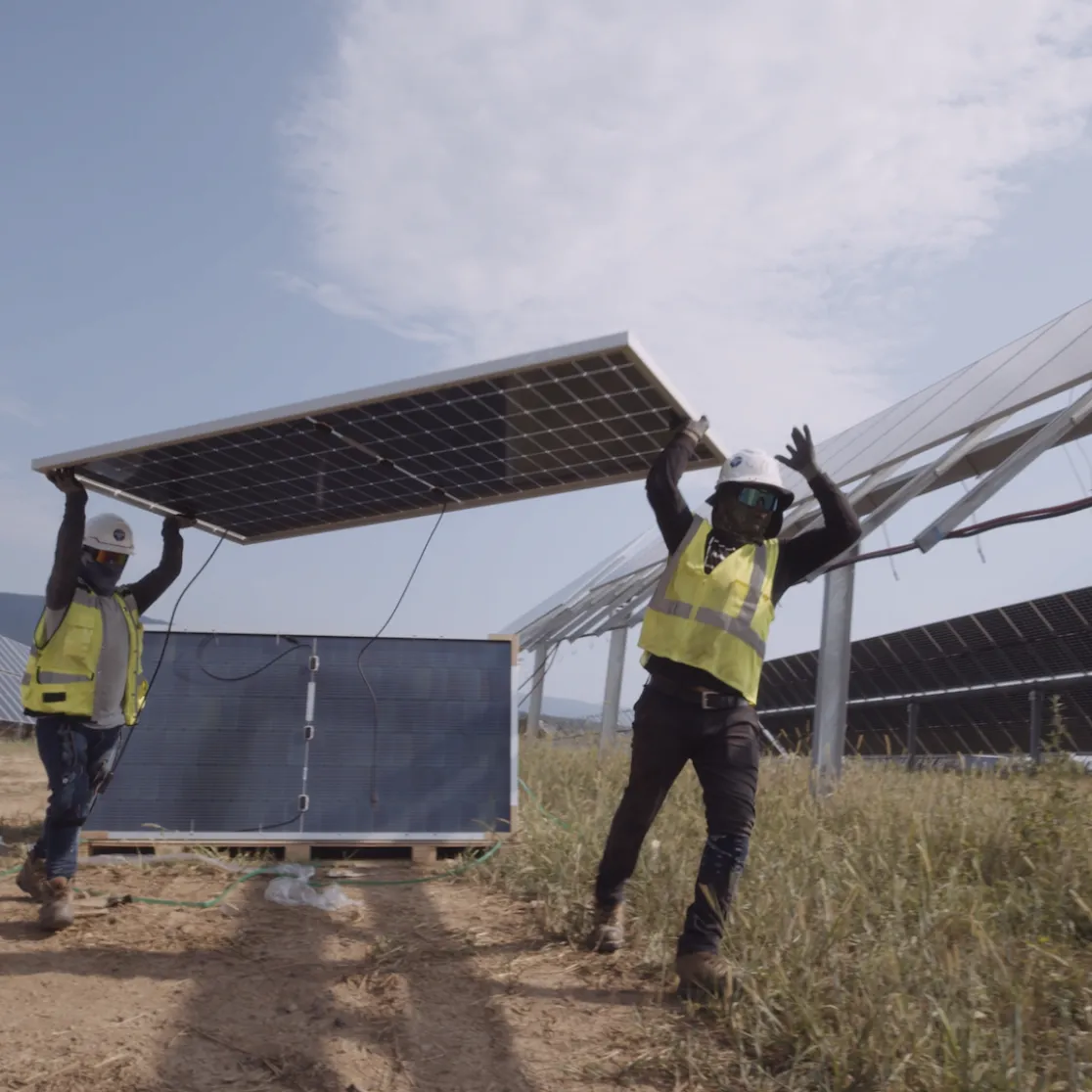 Two workers in safety gear and helmets carry a large solar panel through a solar farm. Rows of solar panels are visible in the background under a partly cloudy sky.