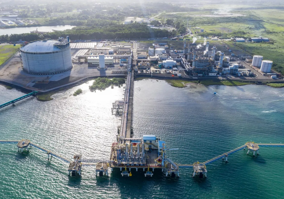 Aerial view of an industrial facility by the water, featuring a large circular storage tank, multiple buildings, and pipelines extending over the water. The facility is surrounded by greenery and has a dock with equipment extending into the sea.