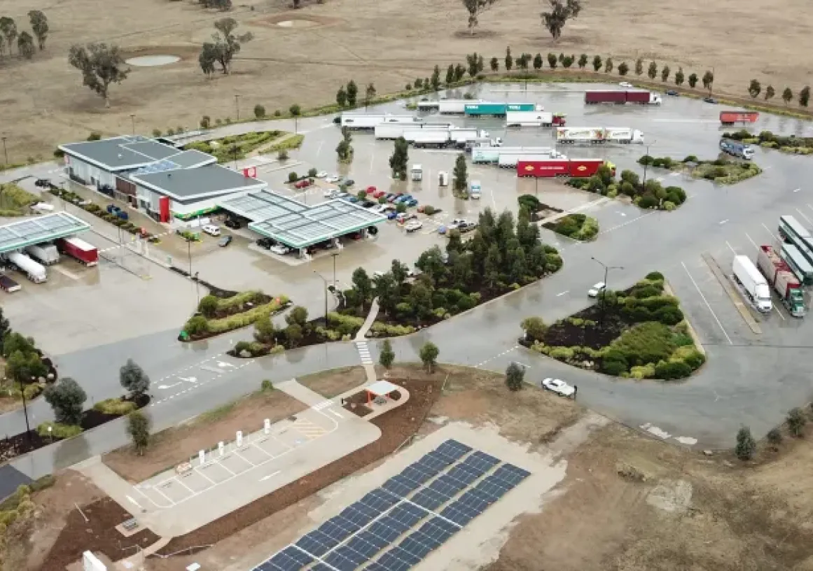 Aerial view of a roadside service station with multiple buildings, parked trucks, and a solar panel installation in a rural area.