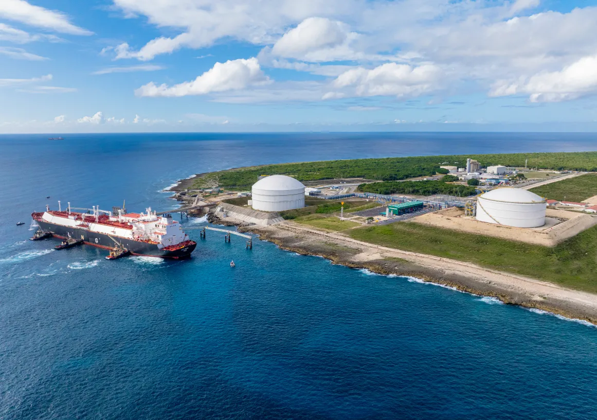 Aerial view of a large tanker ship docked at a coastal LNG terminal with two large white storage tanks. The terminal is situated on a green, grassy landscape next to the ocean under a partly cloudy sky.