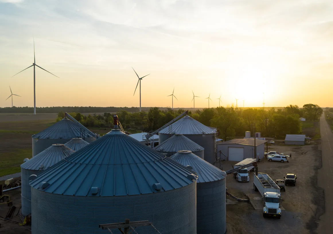 A rural landscape at sunset featuring several large metal silos in the foreground, wind turbines in the background, and a few vehicles parked near a building.