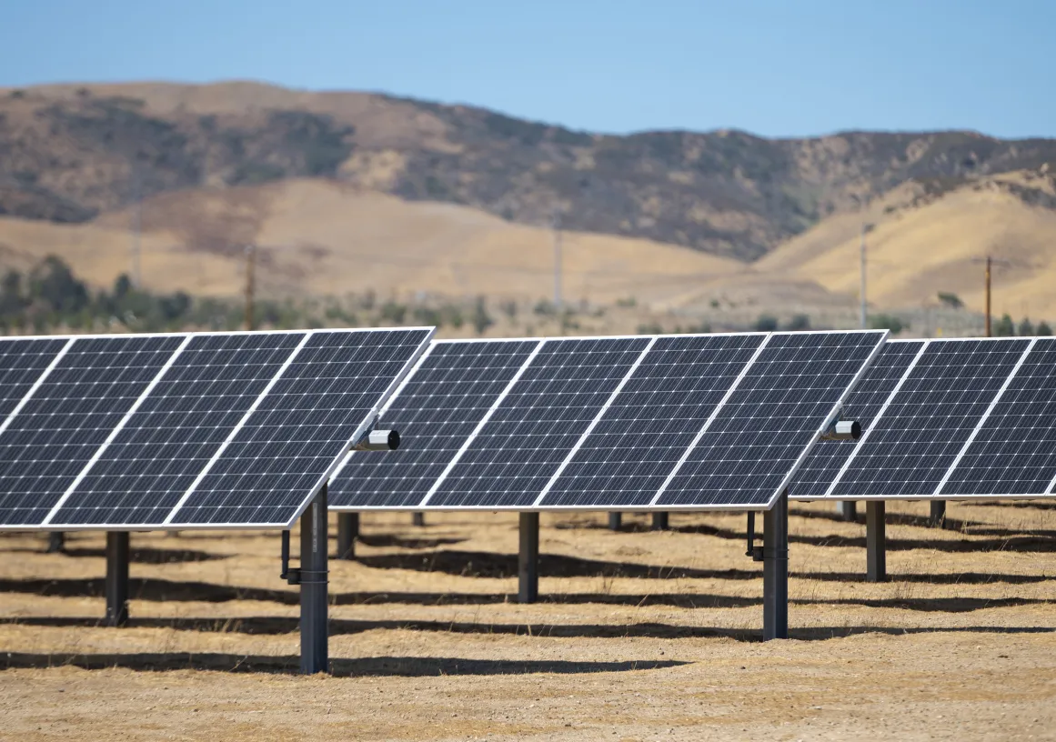 Rows of solar panels installed on a dry, brown field with hills in the background under a clear blue sky.