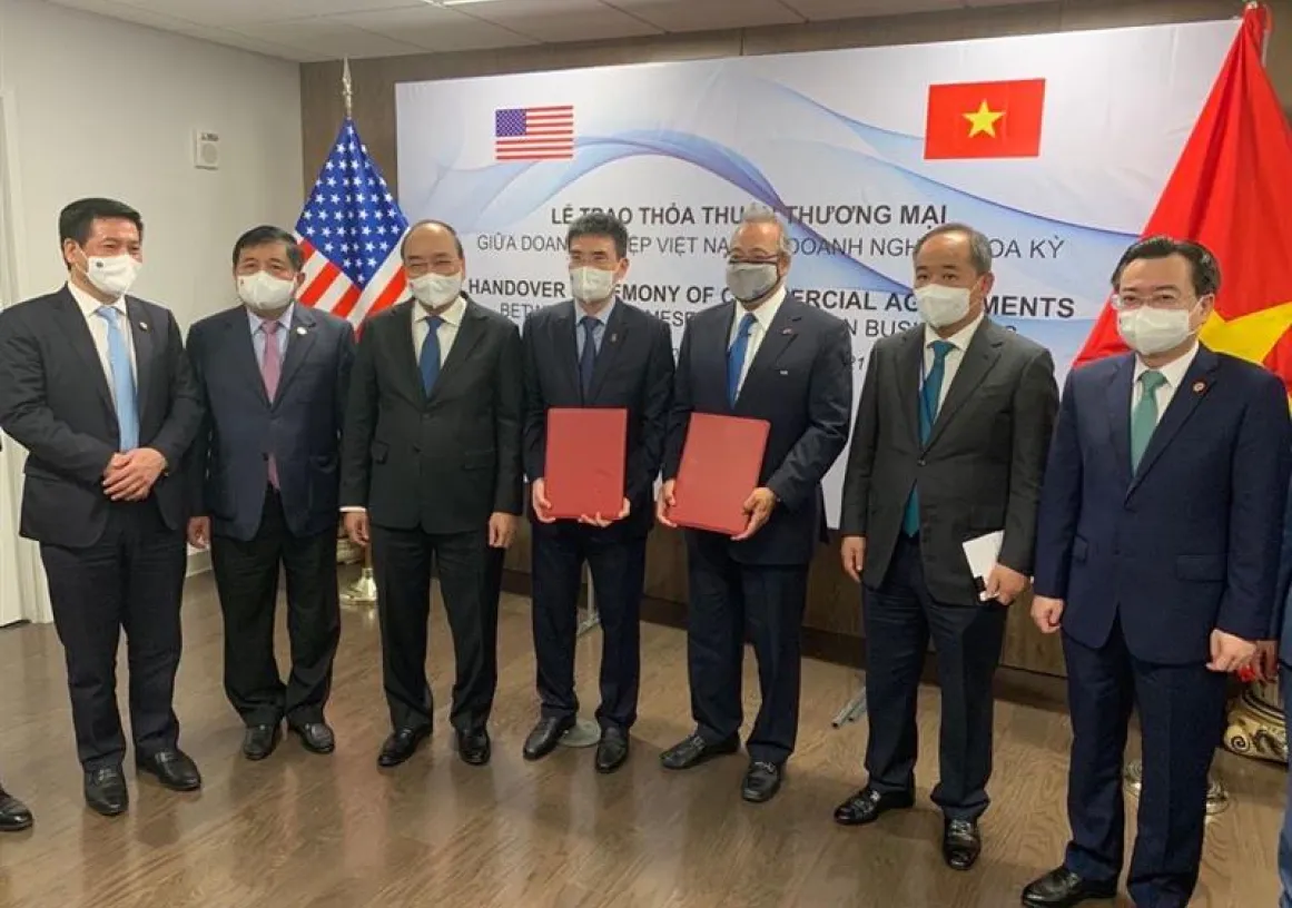 A group of seven men in suits and masks stand in a room with American and Vietnamese flags. Two men in the center hold red folders. A banner behind them reads 'Handover Ceremony of Commercial Agreements' in English and Vietnamese.