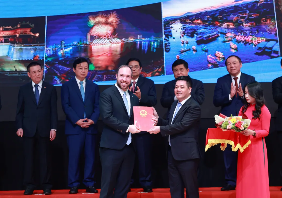 Two men in suits stand on a stage, holding a red document together. Behind them, several other people in suits are clapping. A woman in a red dress holds a floral arrangement on a tray. The background features images of colorful cityscapes and fireworks.