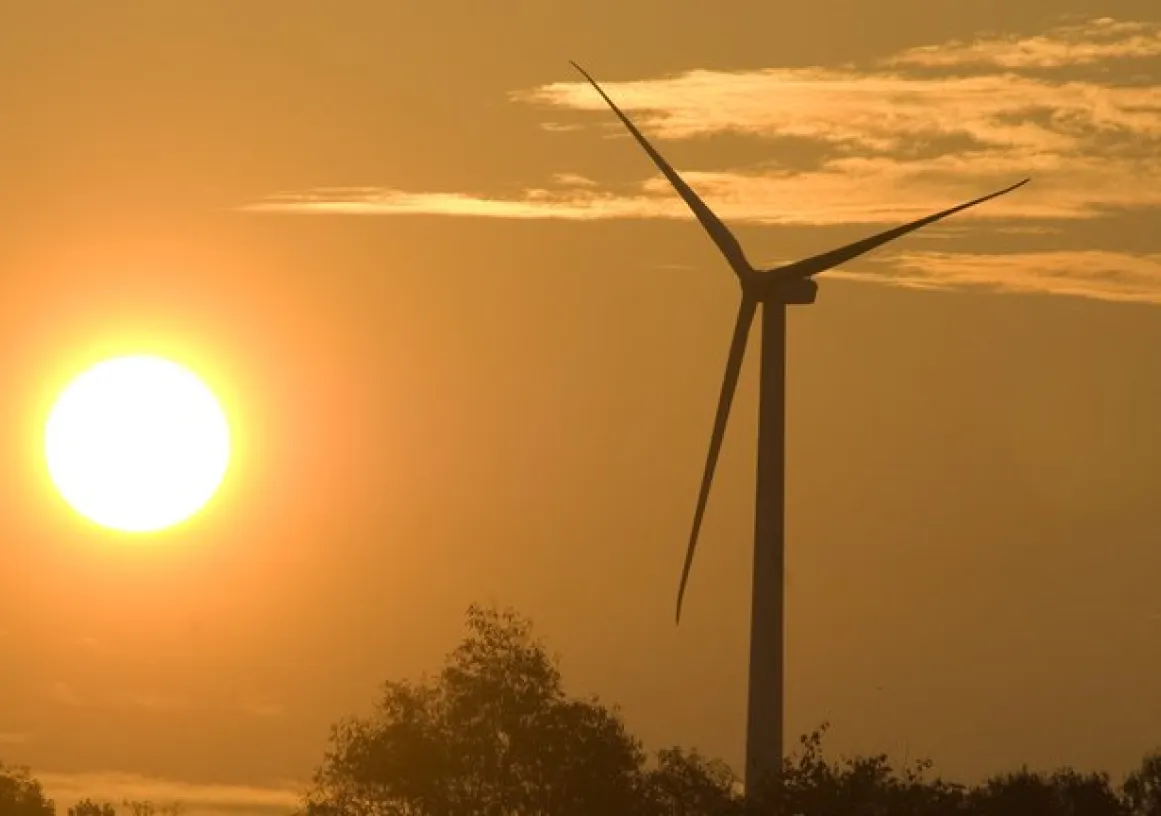 A wind turbine stands tall against a golden sunset sky, with the sun shining brightly near the horizon. Silhouettes of trees are visible at the bottom of the image.