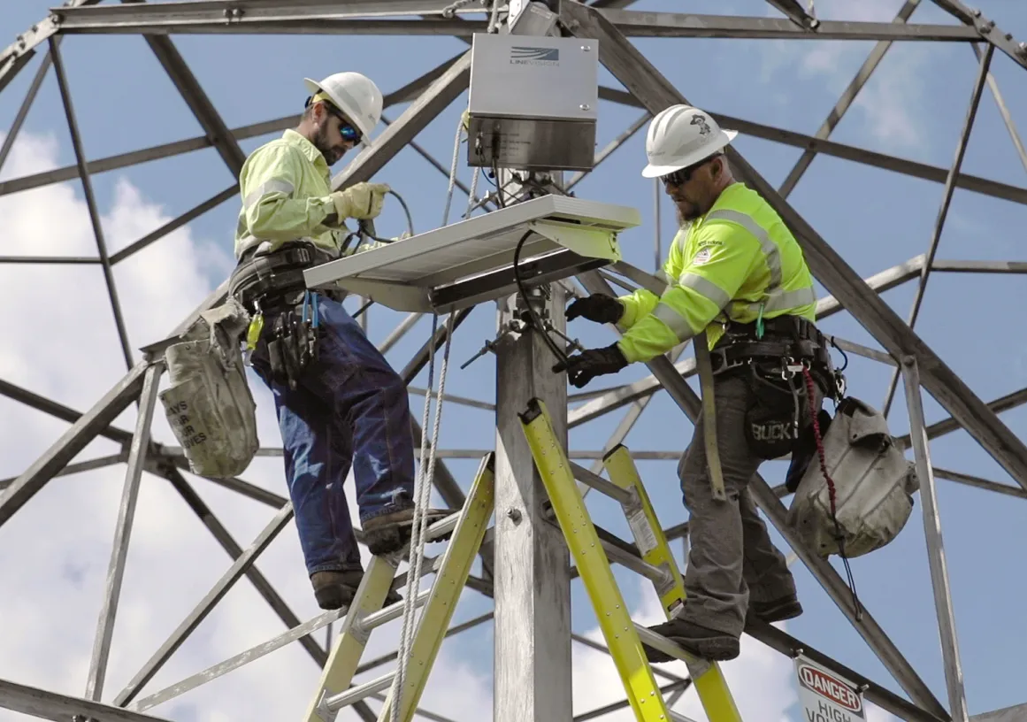 Two workers in safety gear and helmets install equipment on a metal structure. They are on ladders and handling cables, with a clear blue sky in the background.