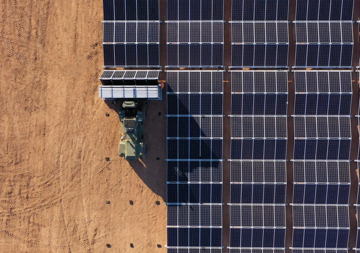 Aerial view of a large solar panel installation on a sandy terrain. A vehicle is parked next to the panels, possibly for maintenance or installation purposes. The panels are aligned in neat rows, casting shadows on the ground.