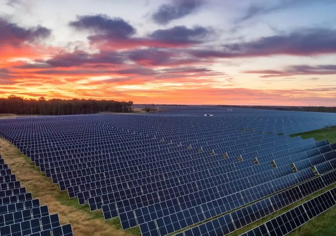 A large field of solar panels under a vibrant sunset sky. The panels are arranged in neat rows, stretching across the landscape. The sky is filled with orange, pink, and purple hues, creating a striking contrast with the dark panels.