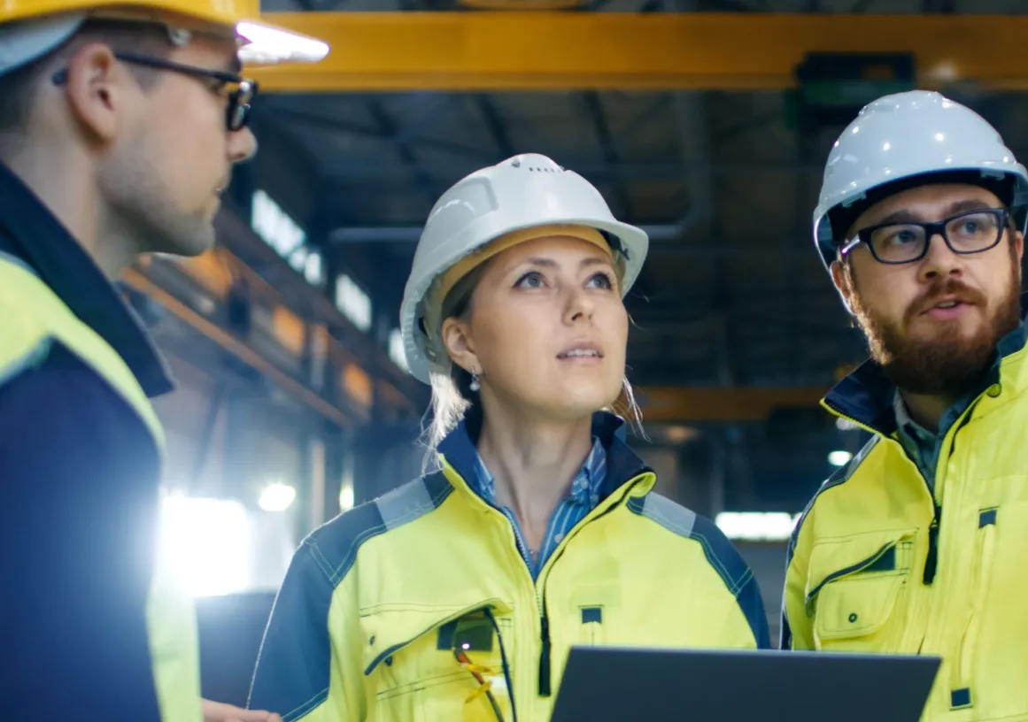 Three construction workers wearing safety helmets and high-visibility jackets stand inside a large industrial facility. One holds a laptop, and they appear to be discussing or inspecting something above.