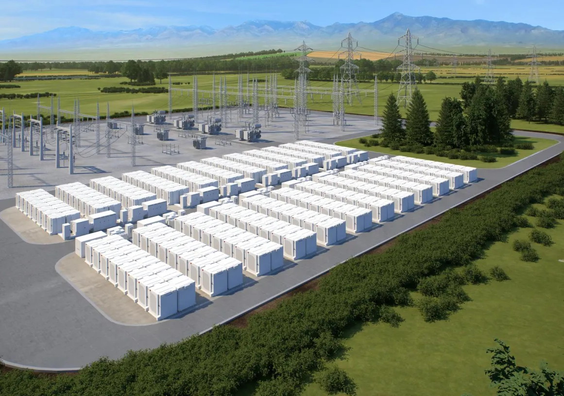 A large outdoor energy storage facility with numerous white battery units arranged in rows. Adjacent to the battery units are electrical substations with metal structures and power lines. The facility is surrounded by green fields and trees, with mountains visible in the background under a clear blue sky.