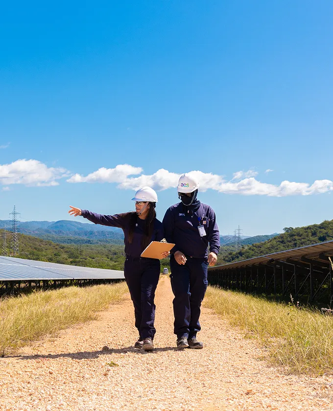 Two engineers wearing helmets walk along a path between solar panels in a solar farm. One points ahead while holding a clipboard. The sky is clear and blue with distant mountains in the background.
