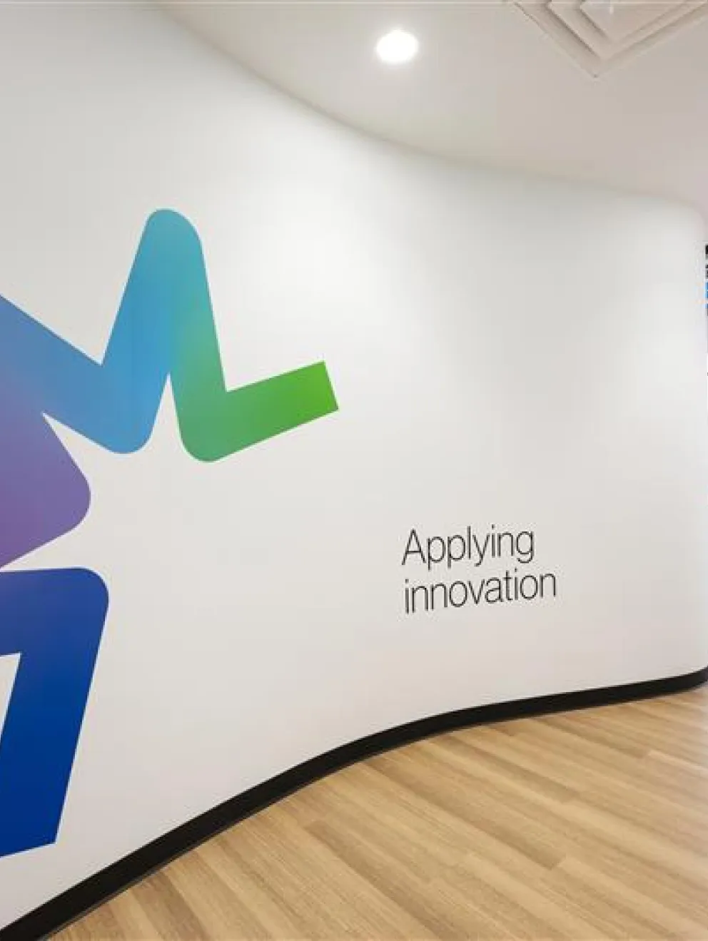 Modern office space with a large abstract logo in blue and green on a white wall, accompanied by the text 'Applying innovation'. The room features wooden flooring and a view of an open area with natural light.
