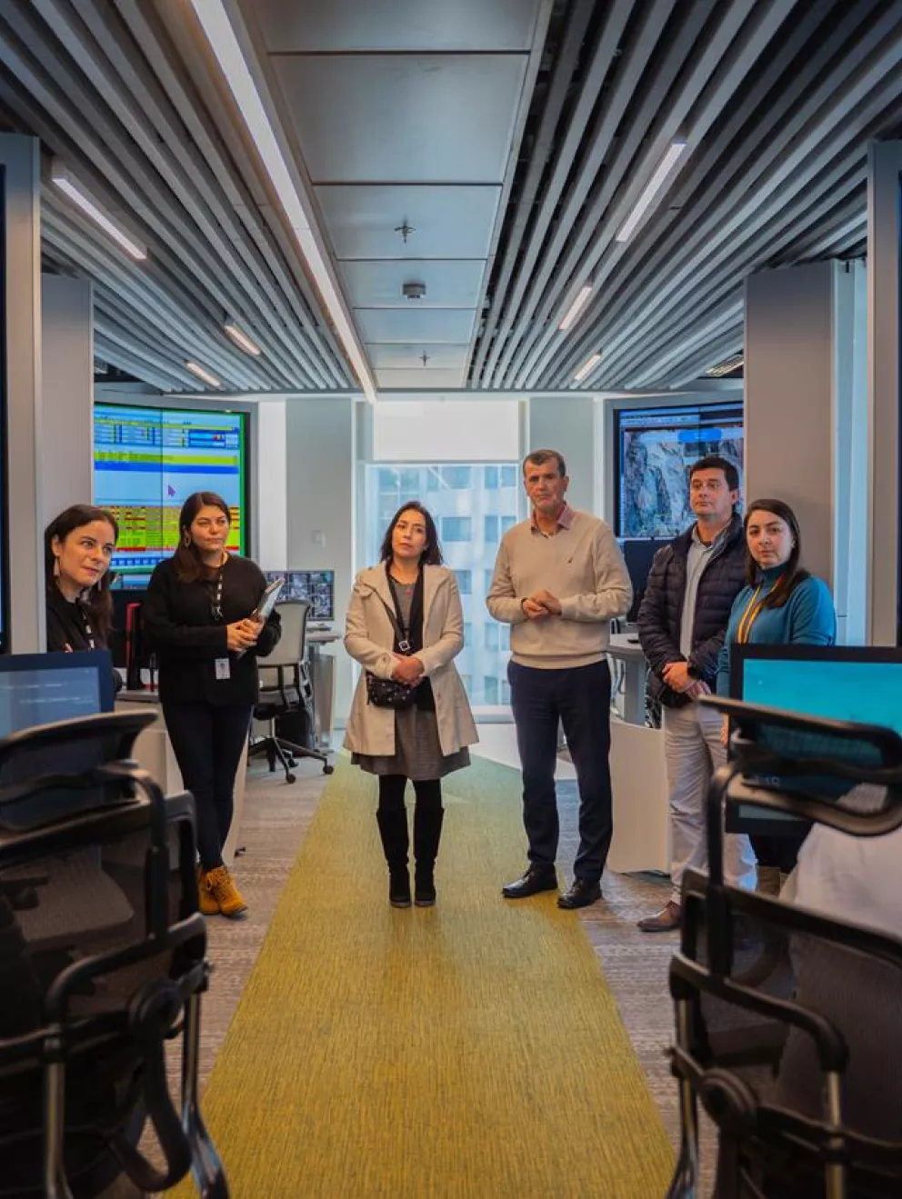 A group of people in an office room with large monitors displaying graphs and data. Some are standing and others are seated at desks with computers.