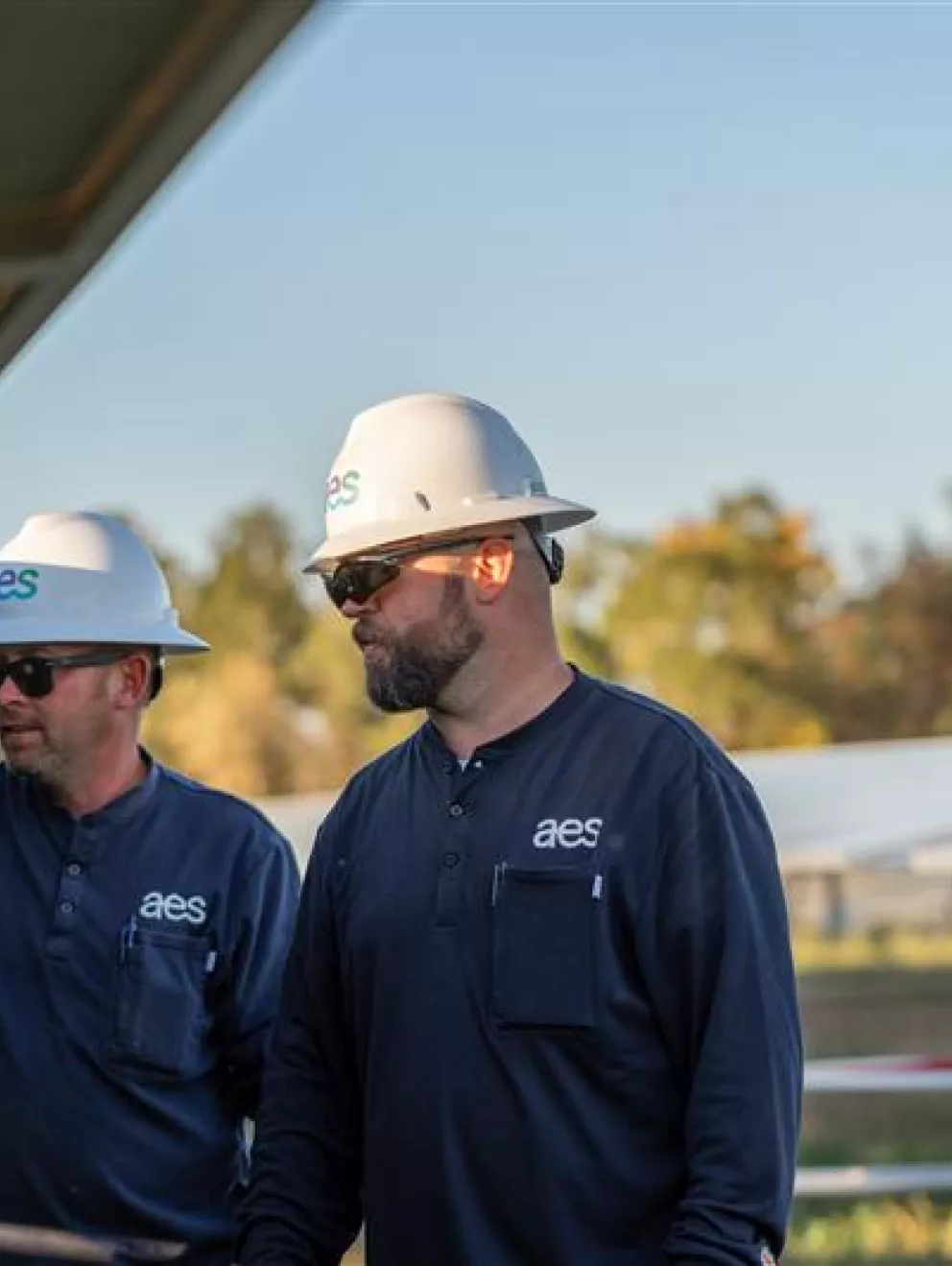 Two workers wearing hard hats and sunglasses inspect solar panels outdoors. They are dressed in dark uniforms with a logo on the chest. The background shows a field with more solar panels and trees under a clear sky.