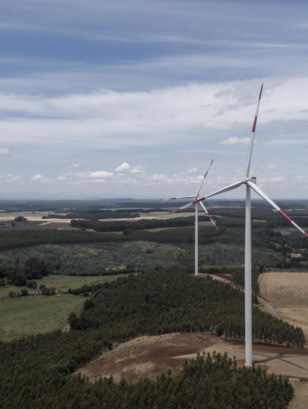 Aerial view of a landscape with two wind turbines on a grassy field surrounded by patches of forest and farmland. The sky is partly cloudy, and the horizon shows distant hills under a blue sky.