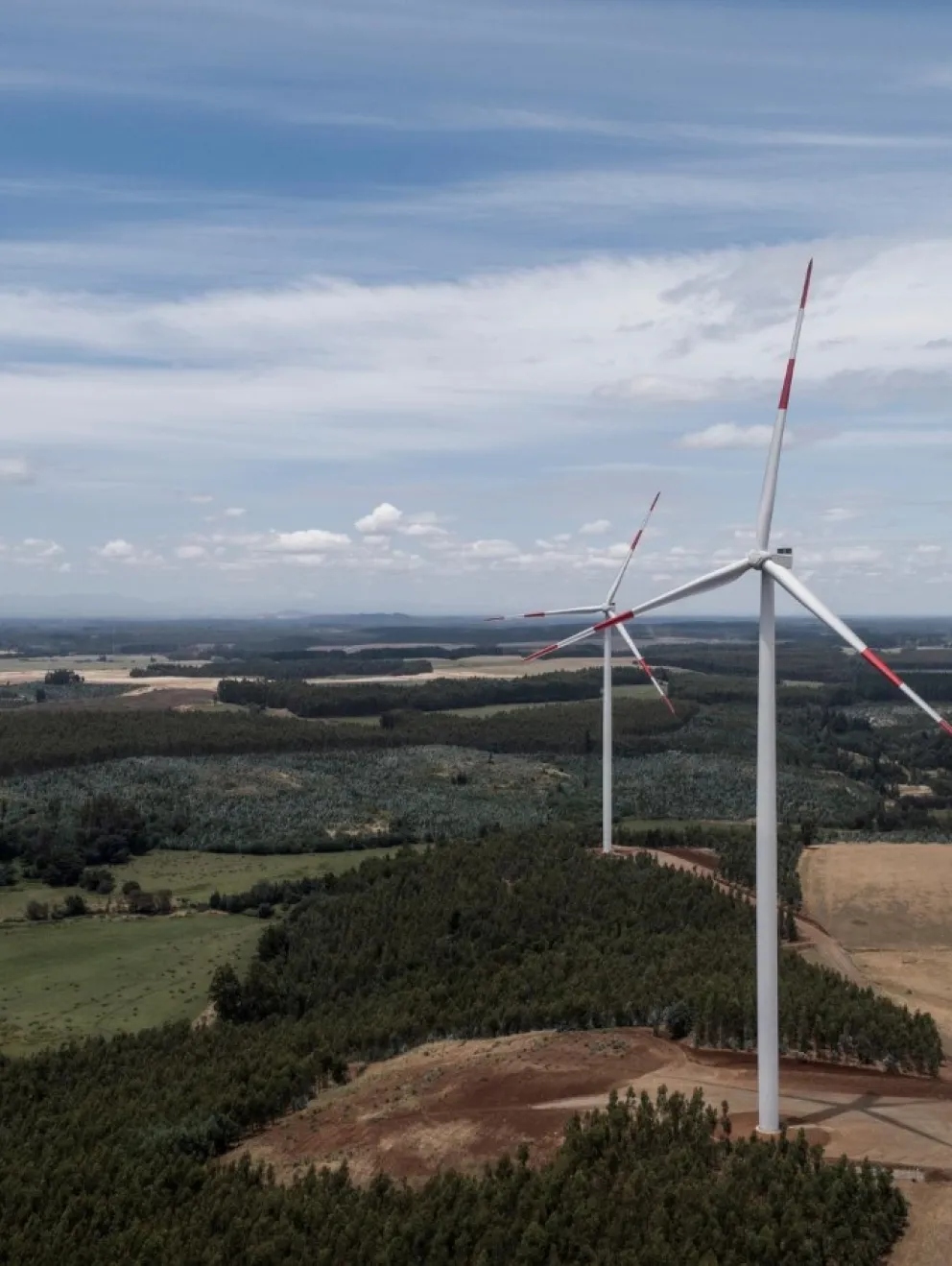 Aerial view of two wind turbines on a vast landscape with green fields and forests under a partly cloudy sky.