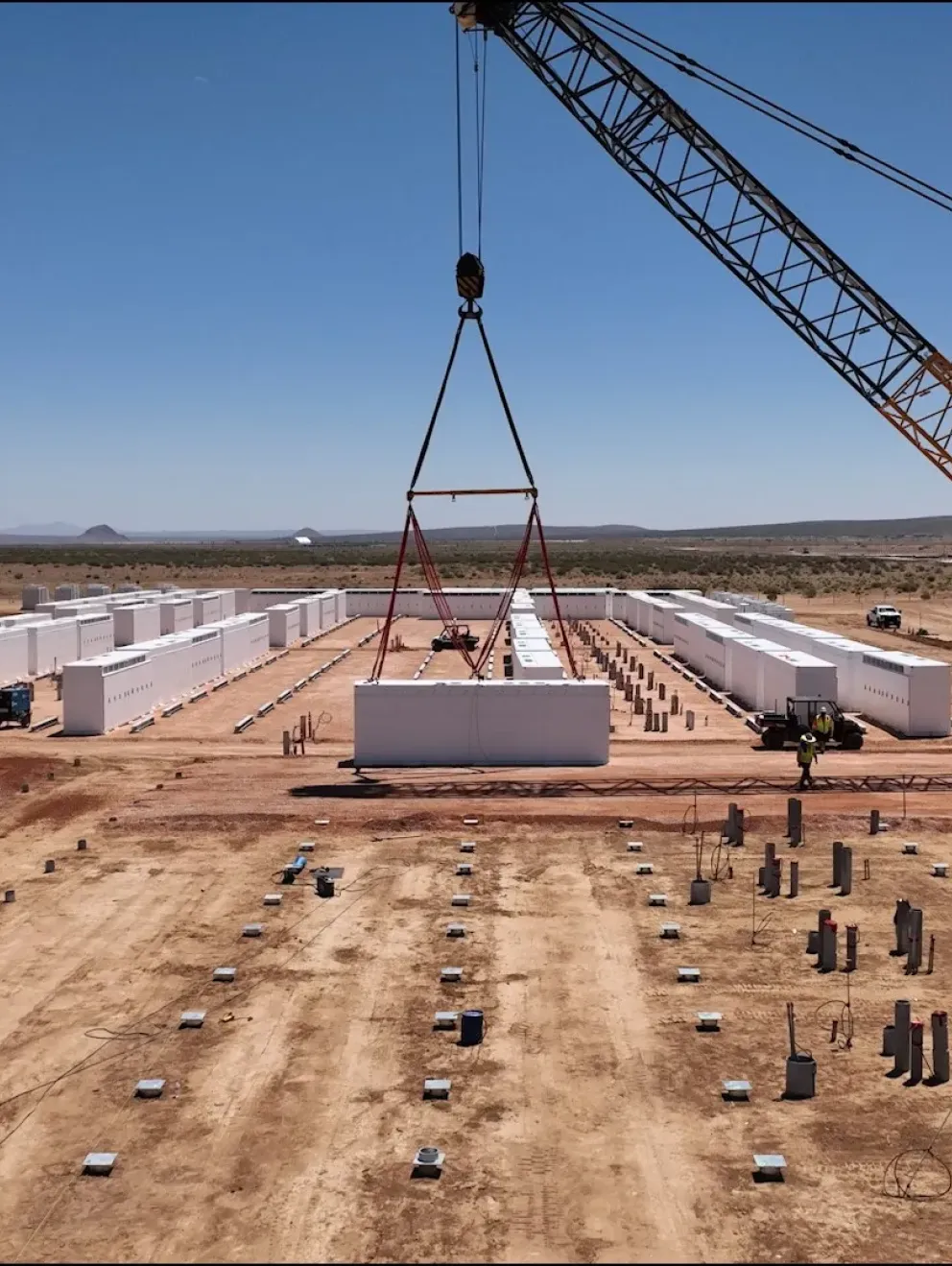 A large crane is lifting a white container in a construction site with several similar containers arranged in rows. The site is in a desert area with mountains in the background. Construction vehicles and workers are visible on the dirt ground.