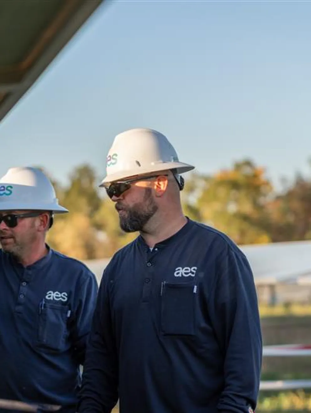 Two workers wearing hard hats and sunglasses inspect solar panels outdoors. They are dressed in dark uniforms with a logo on the chest. The background shows a field with more solar panels and trees under a clear sky.