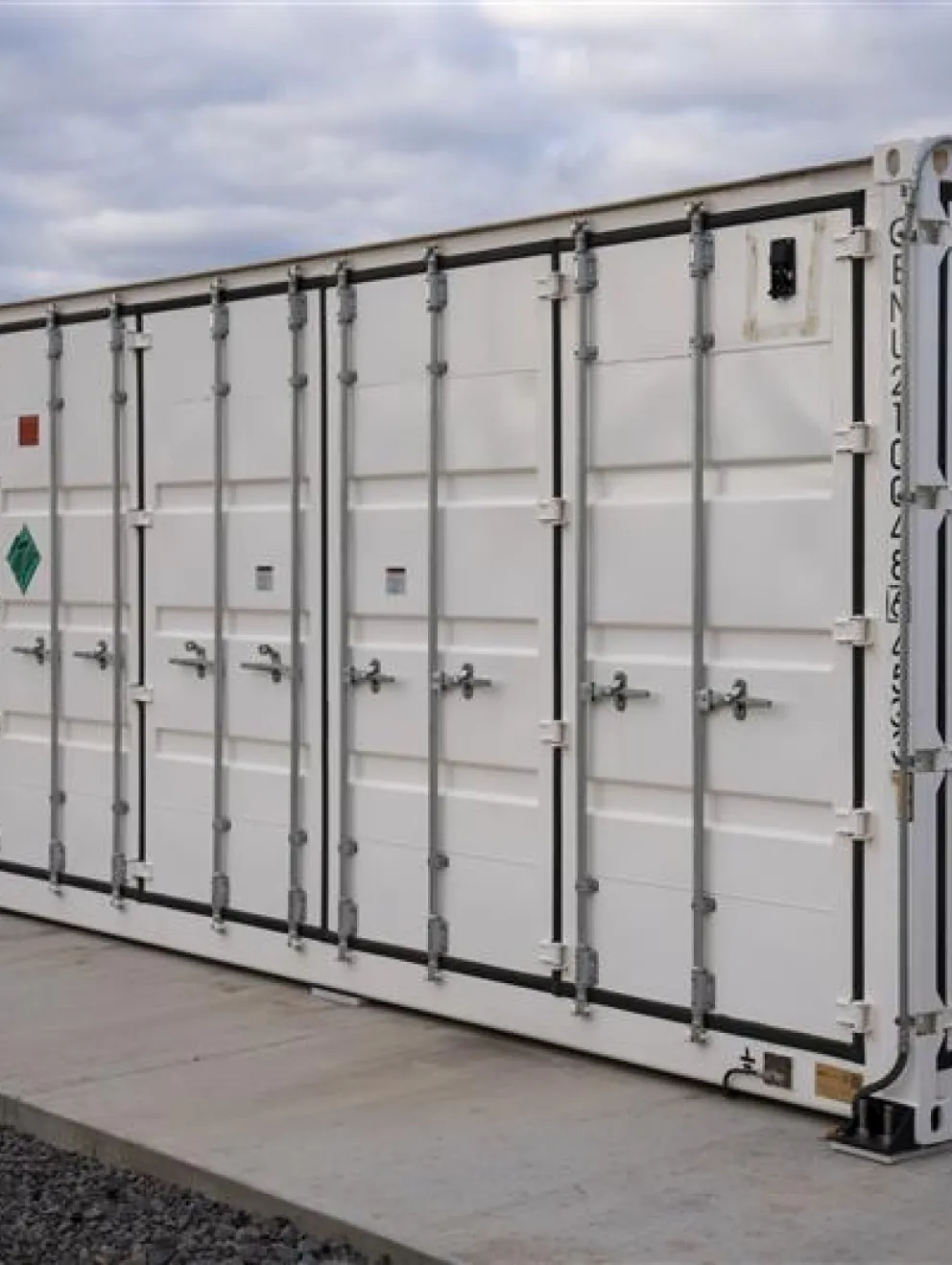 A large white shipping container with multiple doors and hazard symbols is positioned on a concrete platform. The sky is overcast.