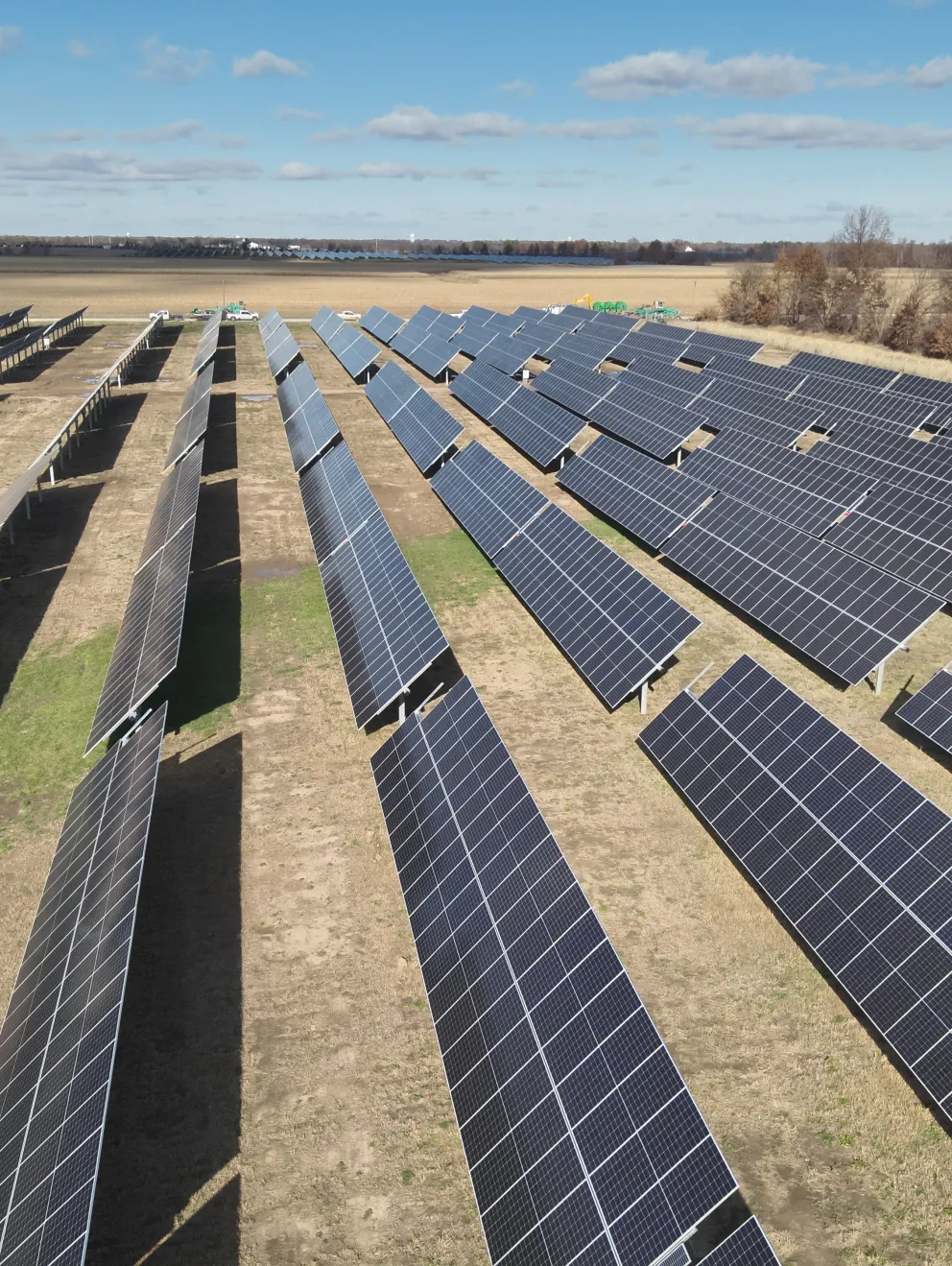 A large field of solar panels arranged in rows under a clear blue sky. The panels are positioned on a flat, grassy terrain with some patches of dirt.