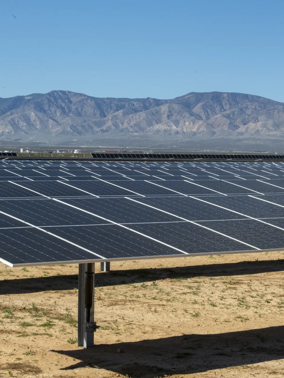 A large solar farm with rows of solar panels stretching across a dry landscape. The panels are aligned in neat rows, reflecting sunlight under a partly cloudy sky. In the background, there are hills and distant mountains.