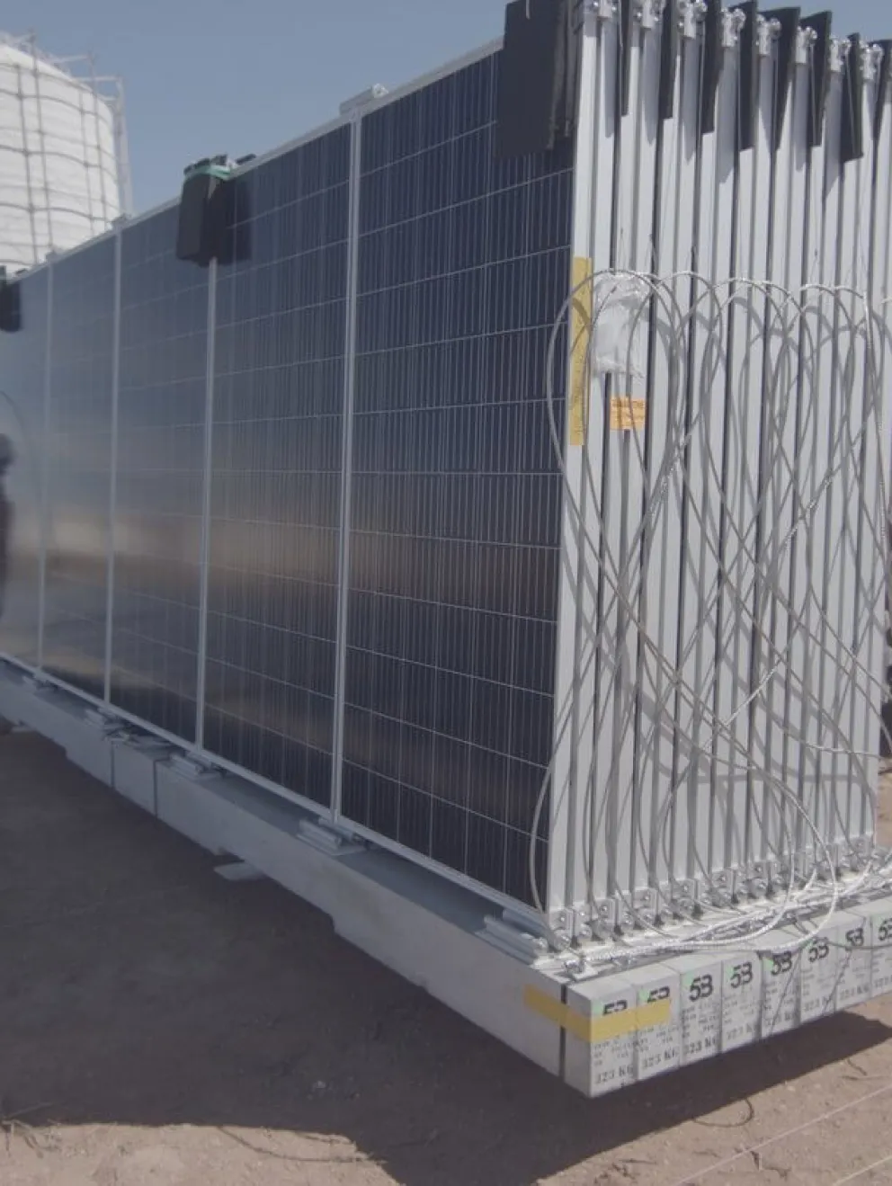 Three workers in safety gear and yellow vests install solar panels on a metal structure in a solar farm. One worker is kneeling, while the others stand on either side, all focusing on securing the panels.