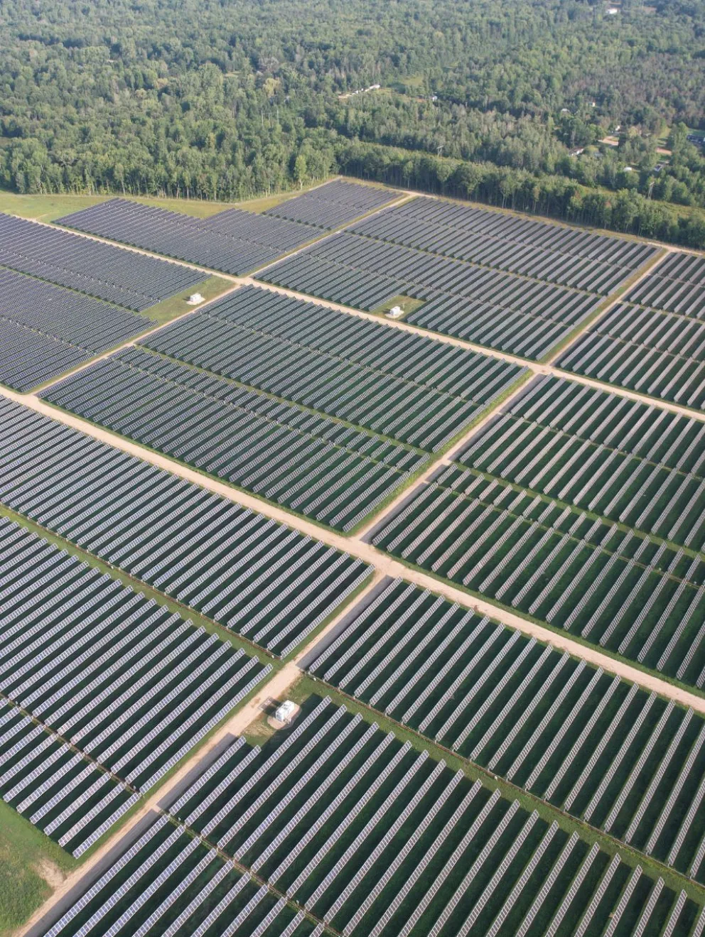 Aerial view of a large solar farm with rows of solar panels arranged in a grid pattern, surrounded by green forested areas.
