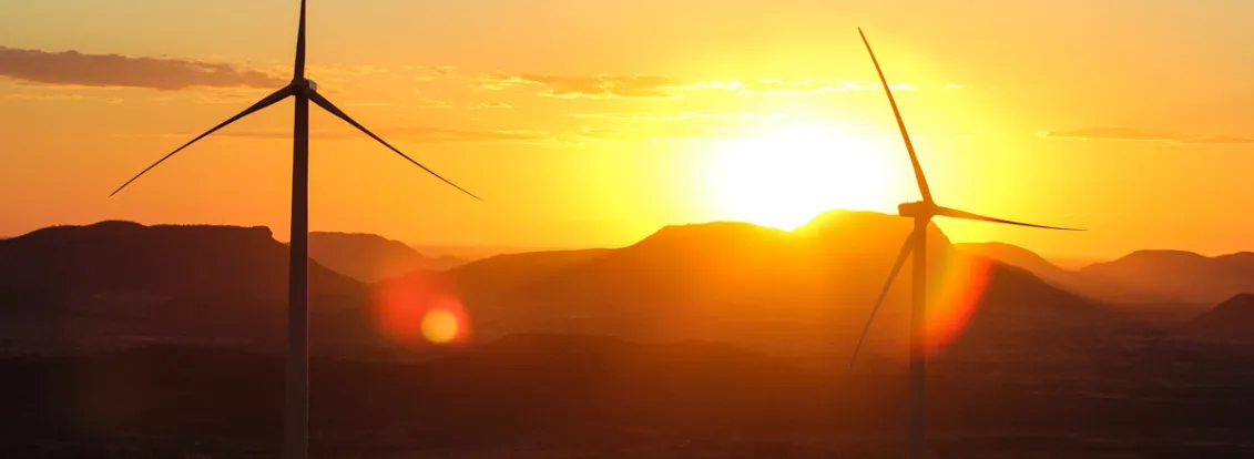 Two wind turbines silhouetted against a vibrant sunset with mountains in the background. The sky is filled with warm hues of orange and yellow, and the sun is partially visible behind the hills.