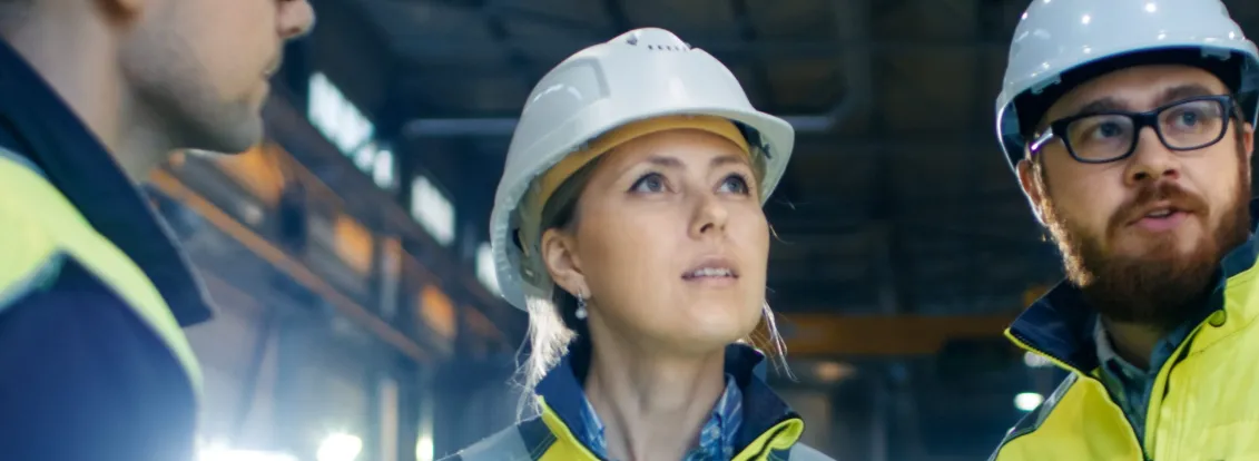Three construction workers wearing safety helmets and high-visibility jackets stand inside a large industrial facility. One holds a laptop, and they appear to be discussing or inspecting something above.