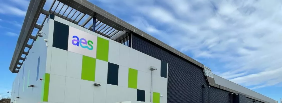 A modern industrial building with a sign displaying 'aes' on the facade. The building has a sleek design with green and dark panels. Several large electrical cabinets are positioned in front of it. The sky is partly cloudy.