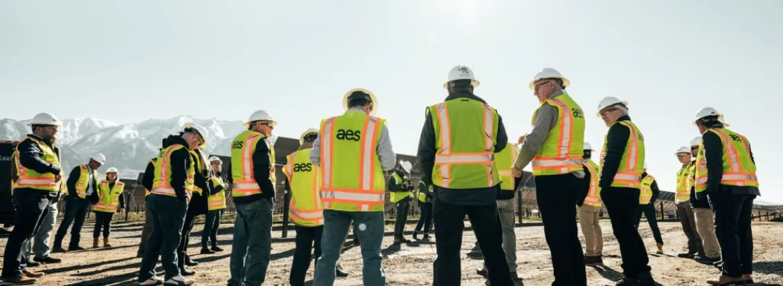 Group of construction workers in high-visibility vests and hard hats standing in a circle on a sunny day, with mountains in the background.