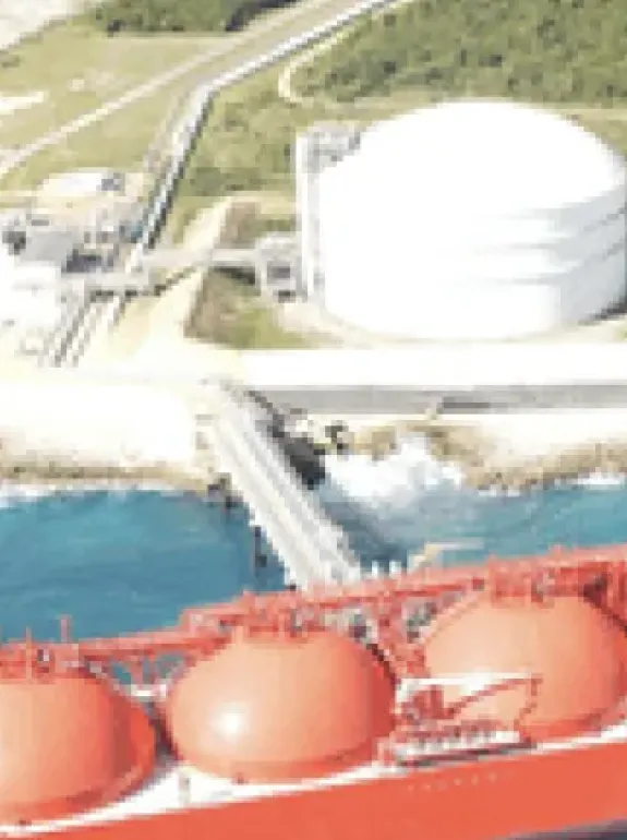 Aerial view of a large red LNG tanker docked near a coastal storage facility with a white spherical tank and surrounding greenery.
