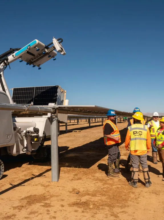 A group of construction workers in safety vests and helmets stand near a solar panel installation site with a large robotic machine labeled 'maximo aes' on a sunny day.